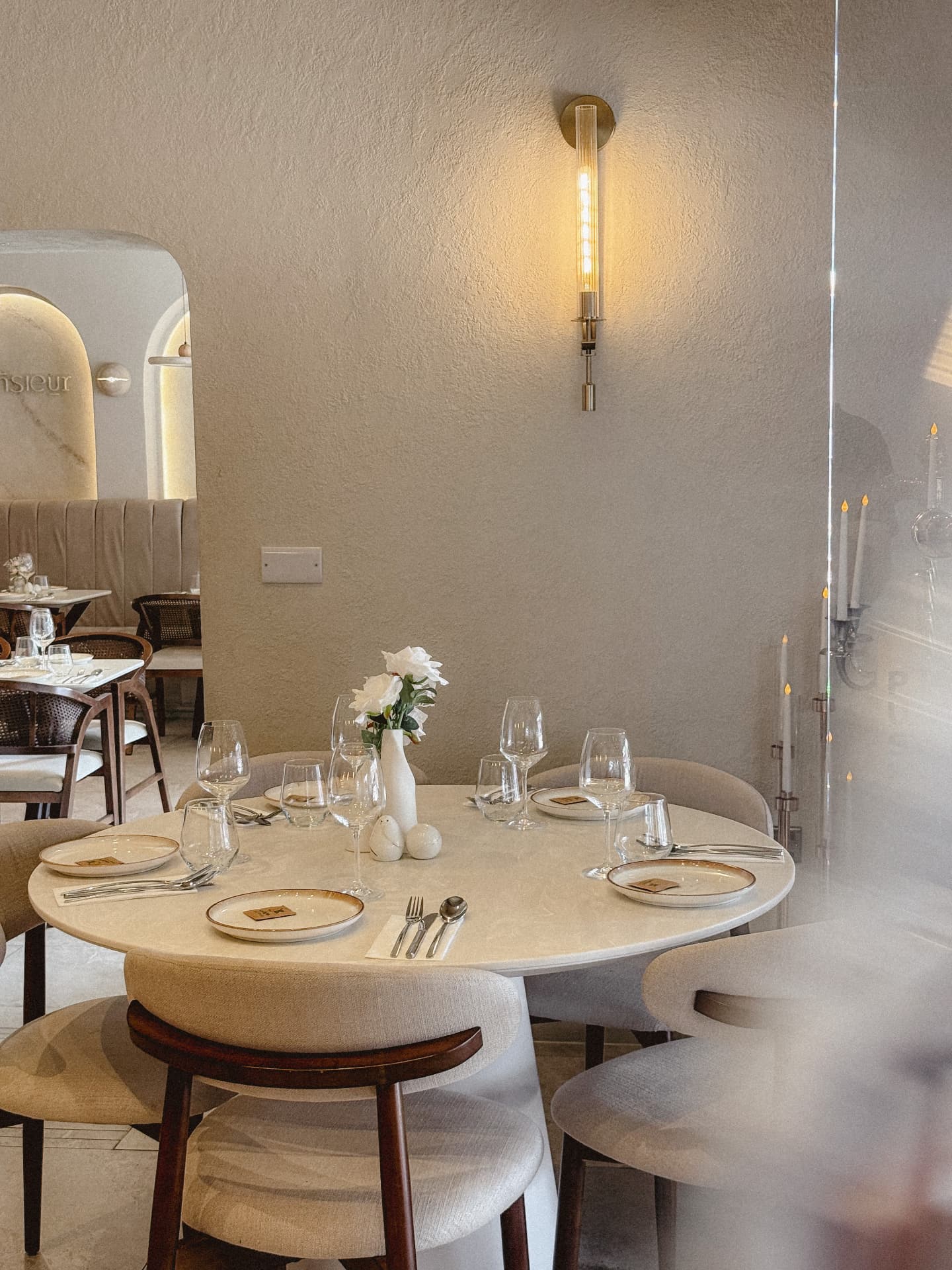 A round cream marble table set beneath a brass wall light, with soft arches and banquettes in the background